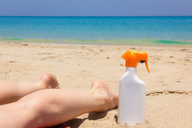 Archivo - Piernas de mujer boca abajo sobre la arena en la playa, botella de spray de loción solar