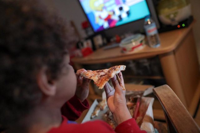 Archivo - Un niño come un trozo de pizza del menú infantil de Telepizza mientras ve la televisión en su casa, tras recoger el menú en un establecimiento de Telepizza.