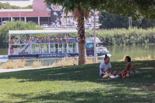 Varias personas se sientan a la sombra junto al río en el parque Magallanes para refrescarse de las altas temperaturas en Sevilla (Andalucía, España). 