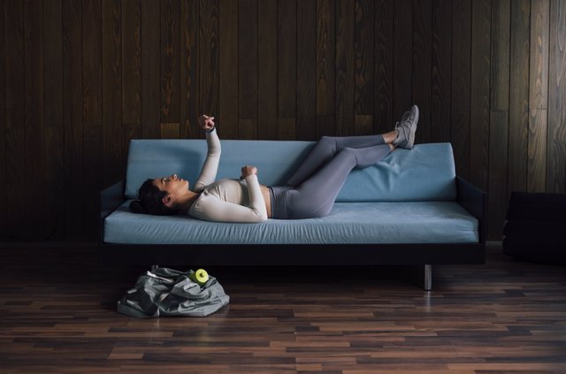 Archivo - Mujer mirando su reloj mientras está acostada en un sofá azul tras volver del gimnasio.
