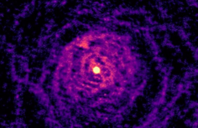 El telescopio ALMA muestra cómo el gas alrededor de la estrella moribunda en W Aquilae ha sido moldeado por interacciones con una estrella compañera similar al Sol. Ambas estrellas están ubicadas en la brillante región central de esta imagen.