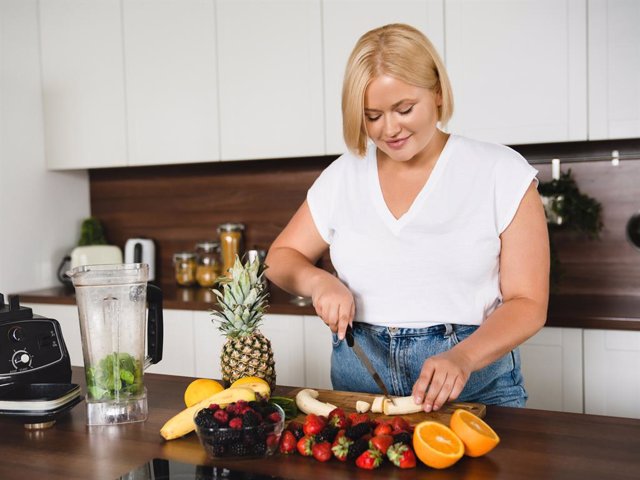 Caucasian plus size plump woman cooking cutting vegetables and fruit for making super food smoothie at home kitchen. Slimming, dieting, shaping concept